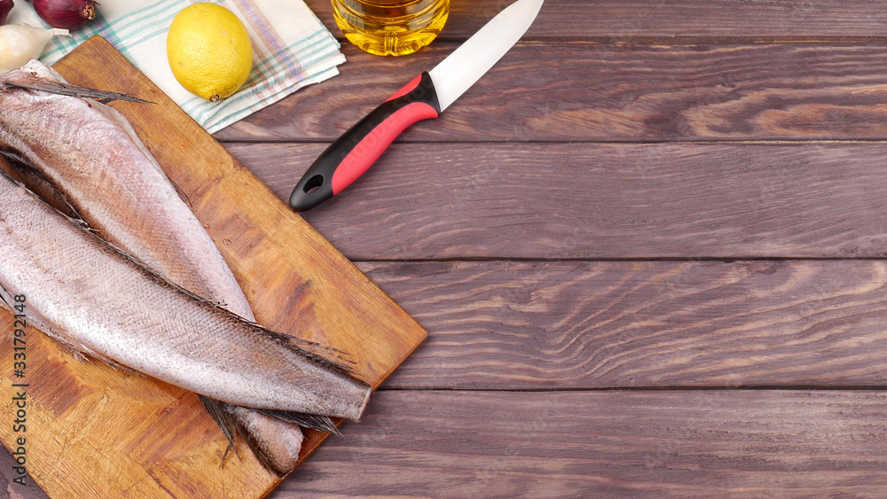 Two carcasses of fresh-frozen hake, pollock fish, on a cutting board on a wooden table. In the background is a knife, lemon, onion and a bottle of sunflower oil. Two carcasses of fresh-frozen hake, pollock fish, on a cutting board on a wooden table. In the background is a knife, lemon, onion and a bottle of sunflower oil.