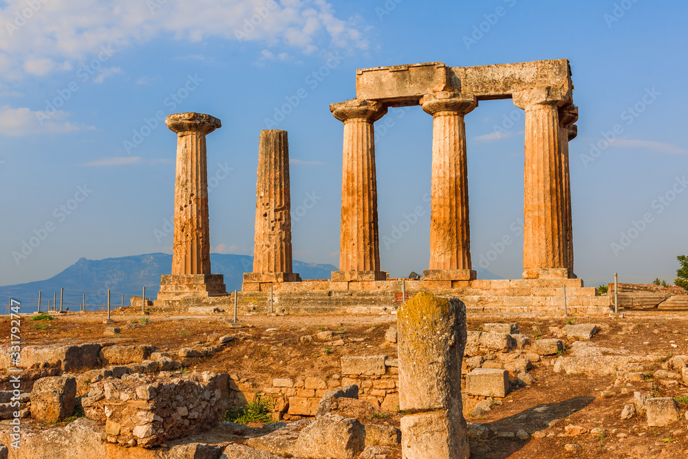 Ruins of Temple of Apollo in Corinth Greece standing up on a hill with remenants of rock walls scattered about under a bright sun with mountians and a blue sky behind