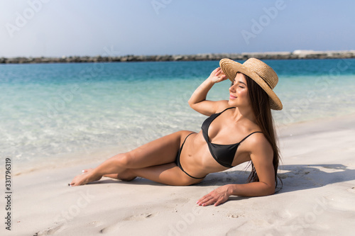 Beautiful woman with perfect body lying down on the beach, wearing stylish hat, tanning on a beach resort, enjoying summer vacation