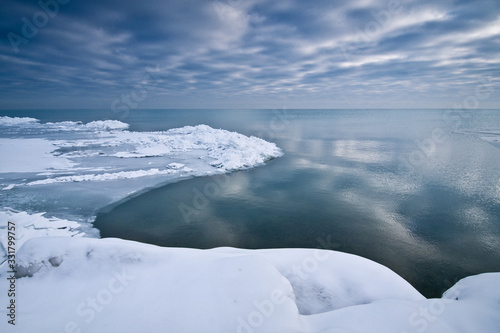 Shelf ice on Lake Michigan shoreline with cloud reflections on calm lake surface.