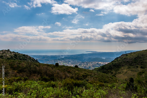 A panoramic view of several towns and their buildings densely covering the coastline behind the low Alps mountains hills with the Mediterranean Sea on the horizon (Provence / Riviera / Côte d'Azur)
