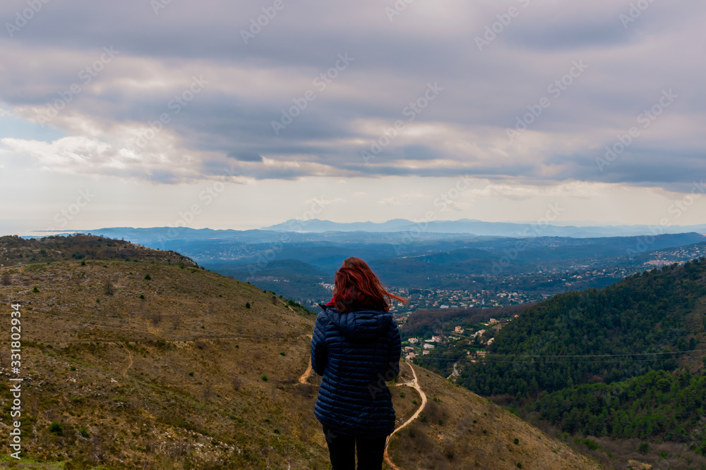 Fototapeta premium A full body back view shot of an unrecognizable young redheaded woman standing enjoying the view of the vast French Alps mountains landscape during a hike in spring