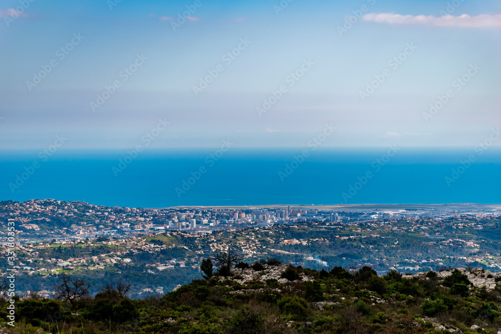 A panoramic view of urban buildings and populated areas densely covering the coastline behind the low Alps mountains hills with the Mediterranean Sea on the horizon (Provence / Riviera / Côte d'Azur)