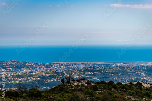 A panoramic view of urban buildings and populated areas densely covering the coastline behind the low Alps mountains hills with the Mediterranean Sea on the horizon (Provence / Riviera / Côte d'Azur)