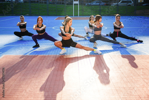 Group of attractive young athletic girls in sportswear doing exercises with squats on the sportground in the green park photo