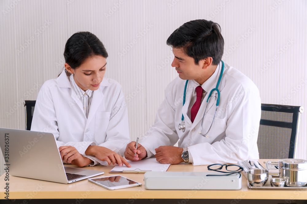 Two doctors wear lab coat and stethoscope discussing patient notes in an office pointing to discuss paperwork together while sitting on the desk with laptops in clinic. Collaboration concept. Two doctors wear lab coat and stethoscope discussing patient notes in an office pointing to discuss paperwork together while sitting on the desk with laptops in clinic. Collaboration concept.