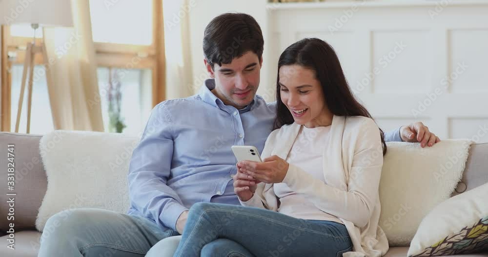 Cheerful young couple having fun watching funny social media videos on phone. Happy millennial man and woman users laughing using cellphone looking at mobile screen relaxing on couch at home together. Cheerful young couple having fun watching funny social media videos on phone. Happy millennial man and woman users laughing using cellphone looking at mobile screen relaxing on couch at home together.