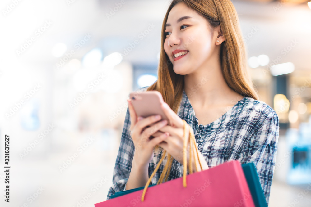 Smiling young Asian woman with shopping colour bags over mall background. using a smart phone shopping online and smiling while standing mall building. lifestyle concept Smiling young Asian woman with shopping colour bags over mall background. using a smart phone shopping online and smiling while standing mall building. lifestyle concept