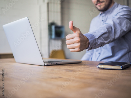 thump up next to laptop and notebook of a happy smiling remote online working man typing and in casual outfit sitting in front of a work desk in an coworking office