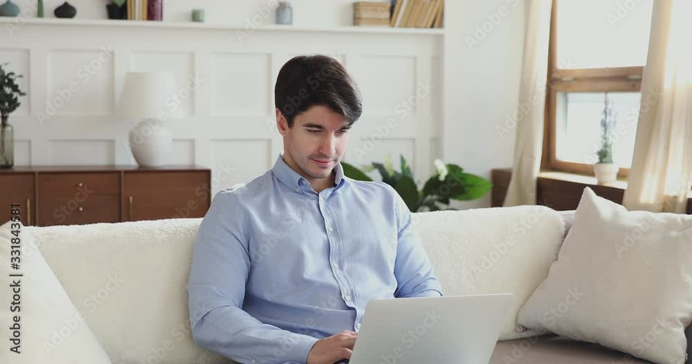 Millennial man using laptop sitting on couch in living room. Young guy browsing internet, surfing web, reading online news, typing on notebook computer modern technology device relaxing at home. Millennial man using laptop sitting on couch in living room. Young guy browsing internet, surfing web, reading online news, typing on notebook computer modern technology device relaxing at home.