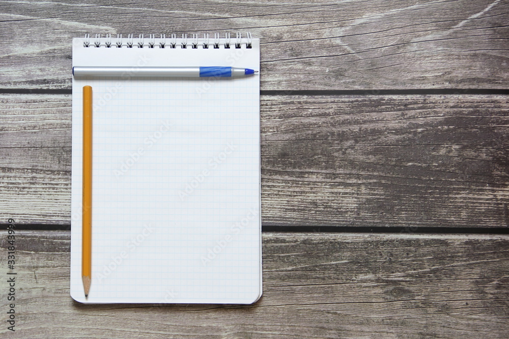 Notepad with a blank white sheet in a checker paper with ball pen and simple graphite pencil lies on the background of wooden boards Notepad with a blank white sheet in a checker paper with ball pen and simple graphite pencil lies on the background of wooden boards