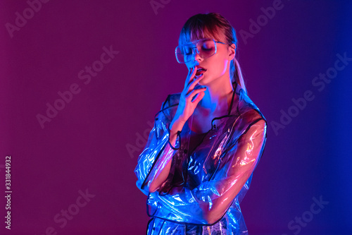 Fashion sensual sexy attractive young woman girl model beauty face wear stylish trendy transparent raincoat eyeglasses eyewear standing in neon light purple blue studio background, copy space.