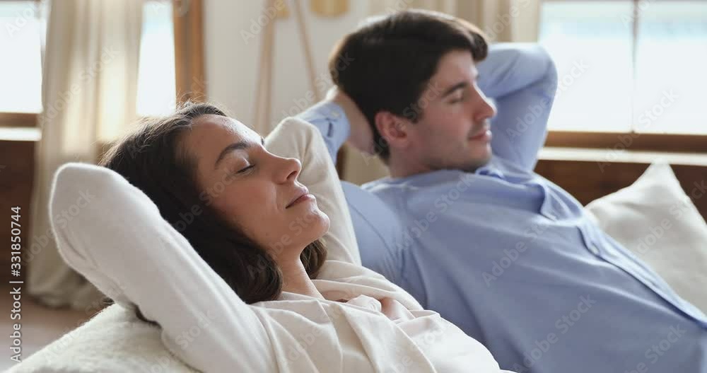 Calm serene young couple relaxing sitting on sofa at home with eyes shut. Smiling lazy husband and wife breathing fresh air, enjoying comfort, no stress in cozy living room holding hands behind head. Calm serene young couple relaxing sitting on sofa at home with eyes shut. Smiling lazy husband and wife breathing fresh air, enjoying comfort, no stress in cozy living room holding hands behind head.