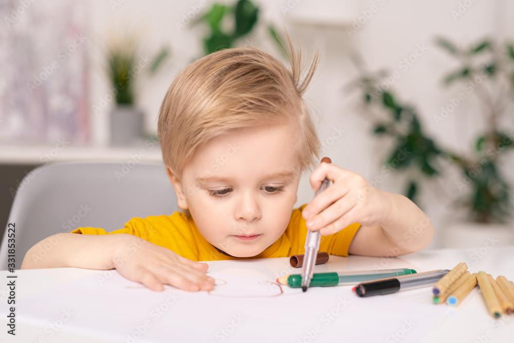Concentrated cute blond-haired boy sitting at table and drawing on paper with colorful felt-tip pens Concentrated cute blond-haired boy sitting at table and drawing on paper with colorful felt-tip pens