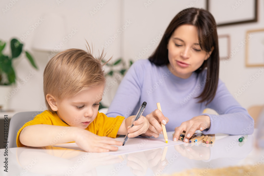 Careful young mother sitting at table and drawing on paper with little son Careful young mother sitting at table and drawing on paper with little son