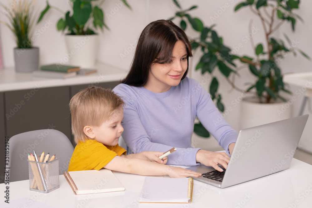 Cute son sitting at table and touching mothers laptop while assisting her with work, young mom working at home Cute son sitting at table and touching mothers laptop while assisting her with work, young mom working at home