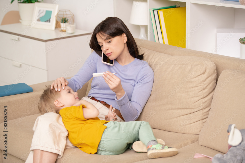 Worried young mother sitting on sofa and checking temperature of son while calling doctor by phone Worried young mother sitting on sofa and checking temperature of son while calling doctor by phone