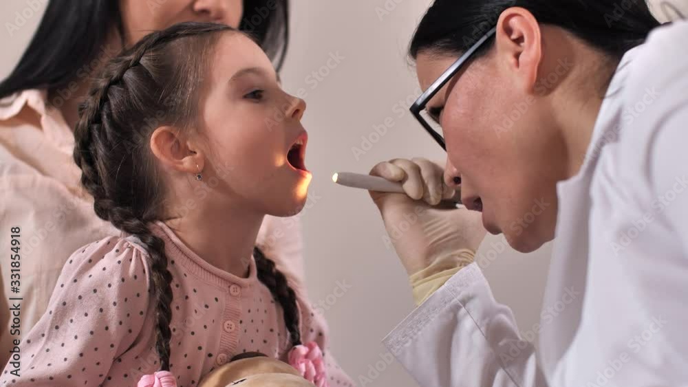 Close-up of cute preschool child opening mouth wide while positive asian female otolaryngologist examining her throat in medical clinic. Sick girl sitting on mom's knees during medical examination Close-up of cute preschool child opening mouth wide while positive asian female otolaryngologist examining her throat in medical clinic. Sick girl sitting on mom's knees during medical examination