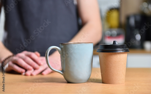 Fototapeta Naklejka Na Ścianę i Meble -  Paper cup of coffee and a mug on the table, against the background of a barista bartender guy. The concept of choice and alternatives