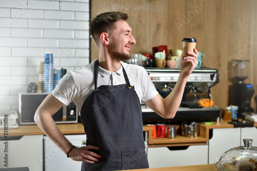 A young barista guy holds a paper cup with coffee in his hands and looks at it. Small business concept