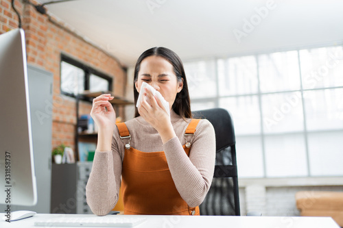  Asian woman sneezes. She uses a tissue to cover her mouth and she is working at home.