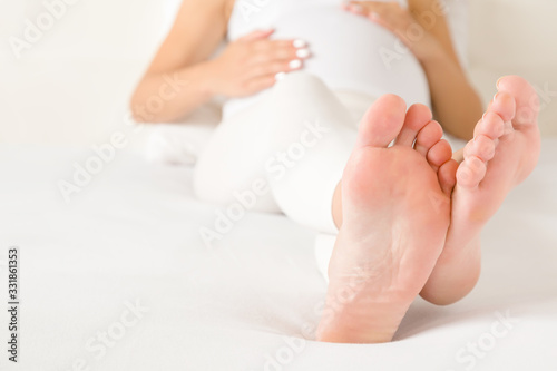 Young woman in white clothes lying in bed and touching belly with hands. Stretched legs. Relaxing feet in pregnancy time. Baby expectation. Barefoot closeup. © fotoduets