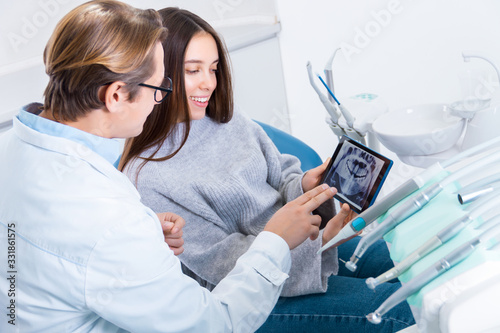 Man doctor is holding tablet computer with panoramic x ray snapshot of jaw on screen and discussing with patient. Young smiling woman is sitting in dental chair in clinic, office. Visit to dentist.