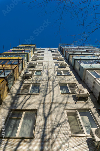 high panel tiled house with many balconies and air conditioning on the facade, shot from below from under trees on a gray cloudy day