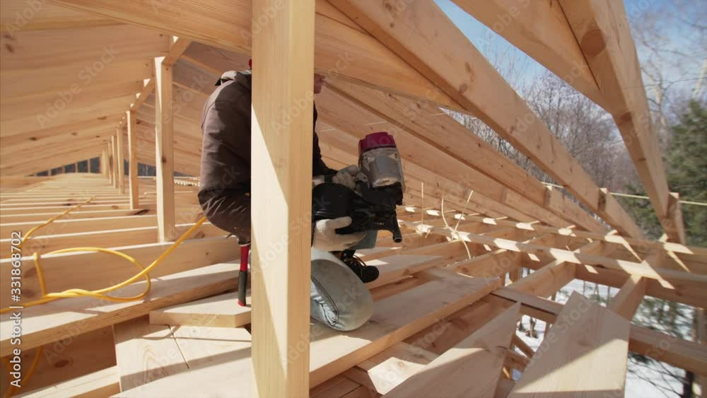 View of builder in red cap nailing wooden planks on the roof with pneumatic nail gun. Frame house under construction View of builder in red cap nailing wooden planks on the roof with pneumatic nail gun. Frame house under construction