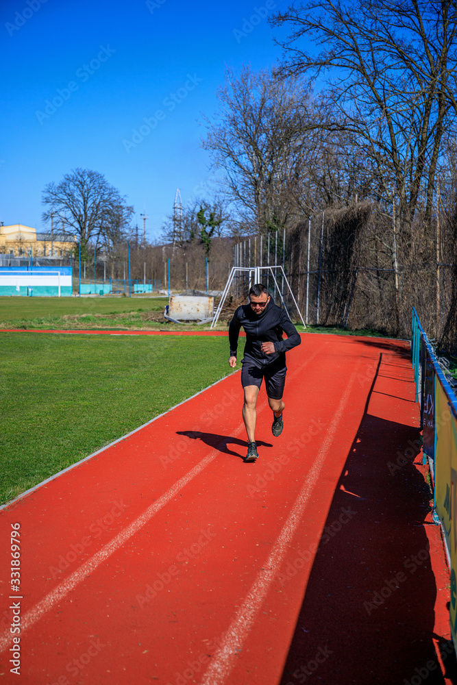 Fit and focused young trandy male athlete running alone along a race track while out training on a sunny day.