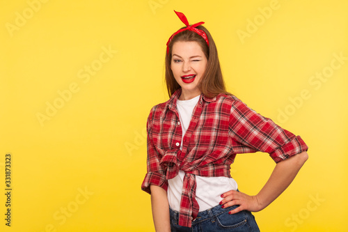 Portrait of cheerful optimistic coquette pinup girl in checkered shirt and headband winking to camera with playful flirty expression