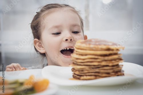 Selected focus. Little girl looking at homemade pancakes with honey on table