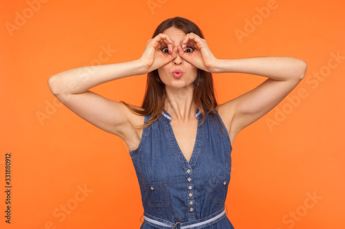 Funny amazed brunette woman making glasses shape with hands, looking through fingers in binoculars gesture and expressing surprise shock, astonishment. indoor studio shot isolated on orange background