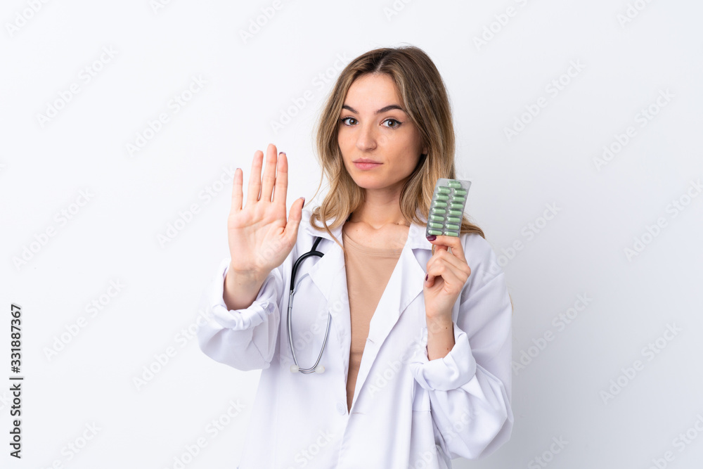 Young woman over isolated white background wearing a doctor gown and holding pills while doing stop sign