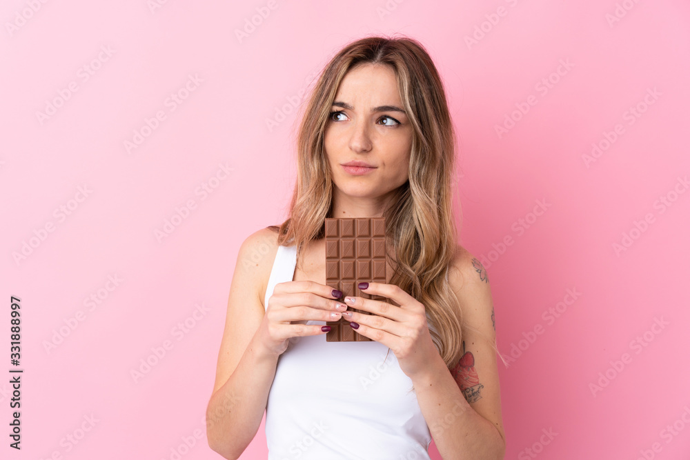Young woman over isolated pink background taking a chocolate tablet and having doubts Young woman over isolated pink background taking a chocolate tablet and having doubts