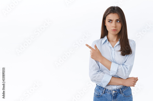 Doubtful and unsure businesswoman looking skeptical with slight disblief, pouting thoughtful while pointing finger left at strangely good offer, hesitating and pondering white background