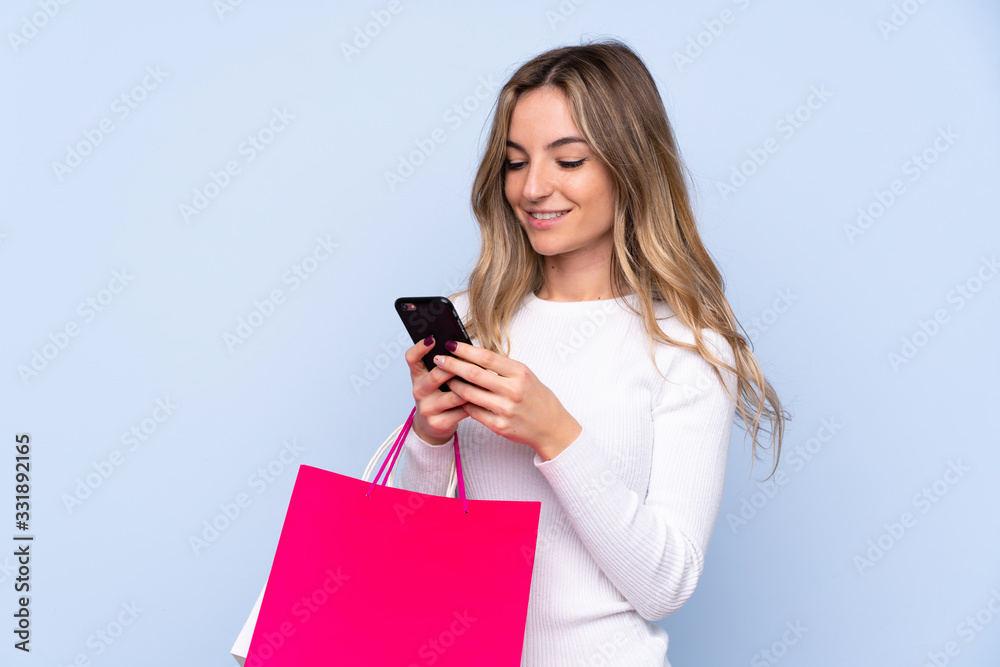 Young woman over isolated blue background holding shopping bags and writing a message with her cell phone to a friend Young woman over isolated blue background holding shopping bags and writing a message with her cell phone to a friend