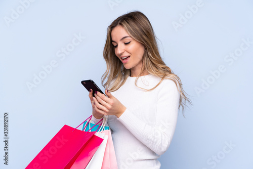 Young woman over isolated blue background holding shopping bags and writing a message with her cell phone to a friend