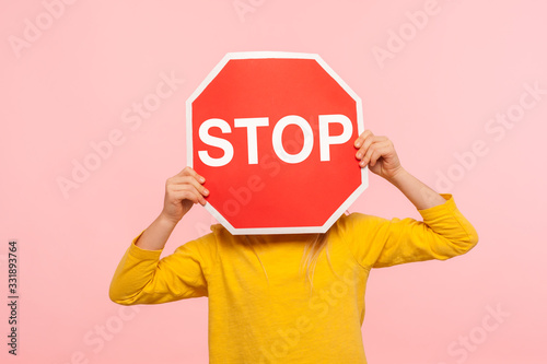 Unrecognizable child in yellow sweatshirt covering face with Stop symbol, anonymous person holding red traffic sign, warning about road safety rules. indoor studio shot isolated on blue background