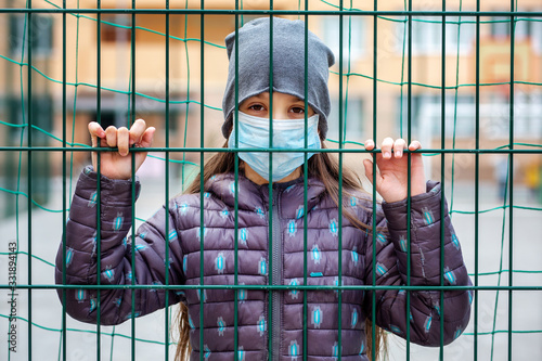 sad young girl in protective medical mask looks at camera through iron mesh in background of building. Concept of coronavirus quarantine in Europe