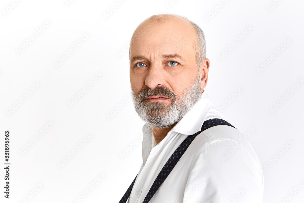 Half profile shot of handsome fashionable senior man with wrinkles, gray beard and blue eyes posing having serious facial expression looking at camera frowning eyebrows wearing elegant clothes