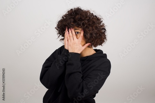Anxiety - a conceptual image of a beautiful young caucasian woman covering her face with her hands and peering out with one eye between her fingers standing indoors. Scared from something or someone.