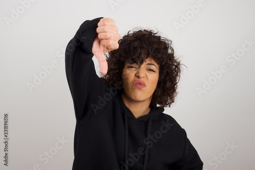 Discontent European woman shows disapproval sign, keeps thumb down, expresses dislike, frowns face in discontent, dressed in white shirt, isolated over gray background. Body language concept. © Jihan