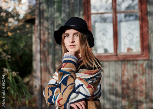 Young blond woman, wearing colorful brown cardigan and black hat, posing in front of old time wooden hut in village in autumn. Pretty hippie girl having fun outdoors on weekend. Three-quarter portrait