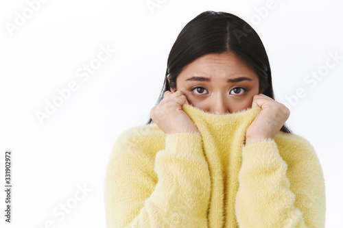 Close-up portrait of timid gloomy cute asian girl, frowning pulling sweater on face and looking camera afraid, trembling fear, being insecure or anxious, standing frightened white background