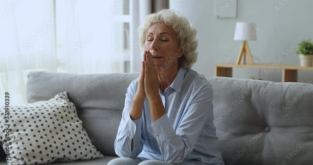 Faithful religious elderly woman sitting on couch, praying god. Mindful hopeful middle aged lady believer joining hands together, saying whispering prayer or grace with closed eyes alone at home. Faithful religious elderly woman sitting on couch, praying god. Mindful hopeful middle aged lady believer joining hands together, saying whispering prayer or grace with closed eyes alone at home.