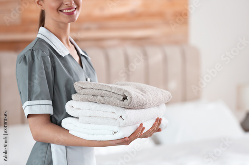 Young chambermaid holding stack of fresh towels in bedroom, closeup