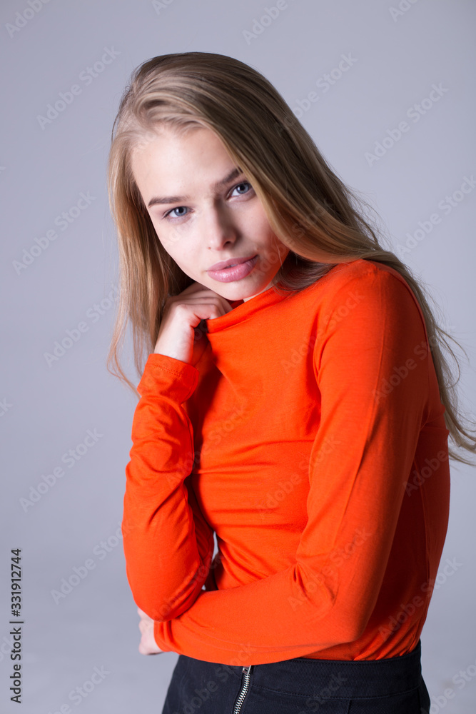 Nice portrait of well-dressed fashionable girl in orange sweatshirt. Lovely lady posing indoors on gray background. Blonde model with serious look. Fashion and lifestyle concept Nice portrait of well-dressed fashionable girl in orange sweatshirt. Lovely lady posing indoors on gray background. Blonde model with serious look. Fashion and lifestyle concept