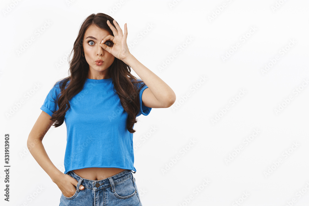 Impressed and curious cute curly-haired feminine brunette woman in basic blue t-shirt, showing okay over eye, folding lips amazed, say wow to interesting thing, intrigued of what found Impressed and curious cute curly-haired feminine brunette woman in basic blue t-shirt, showing okay over eye, folding lips amazed, say wow to interesting thing, intrigued of what found