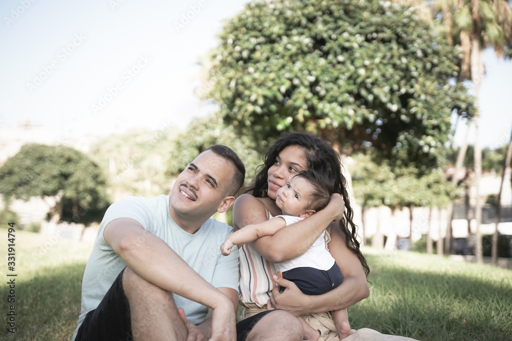 Fototapeta premium Mother, father and baby sitting on the grass of the public park. Happy young family spending time together outside - Image